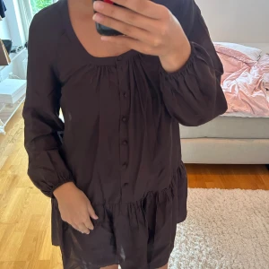 Brun kort klänning med knappar - Säljer en brun kort klänning med långa ärmar och rund halsringning. Klänningen har knappar framtill och ett lätt rynkat parti vid midjan som ger en avslappnad och flowy känsla. Perfekt för dig som gillar enkel och stilren design.