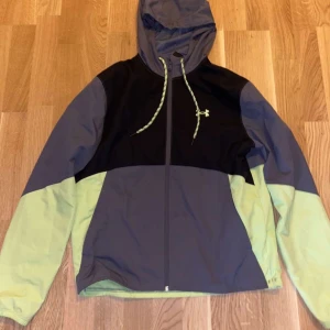Under Armour vindjacka med huva - Snygg vindjacka från Under Armour i svart, grått och ljusgrönt. Jackan har huva med snörning, dragkedja framtill och logga på bröstet. Perfekt för blåsiga dagar och sportiga outfits. Långärmad och tillverkad i lätt polyester.