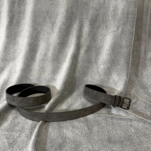 Mörkgrått mockabälte med metallspänne - Stilrent mörkgrått bälte i mocka med klassiskt svart metallspänne. Bältet har en enkel och clean design utan mönster, perfekt för att lyfta vilken outfit som helst. Bältet är rakt och har flera hål för justerbar passform. Längd ~110 cm 