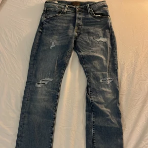 Jack & Jones blå slitna jeans - Snygga blå jeans från Jack & Jones med slitningar på låren och knäna. Klassisk femficksmodell med raka ben och normal passform. Jeansen har ljus tvätt och detaljerade bakfickor med läderpatch i midjan. Modellen är slim/glenn