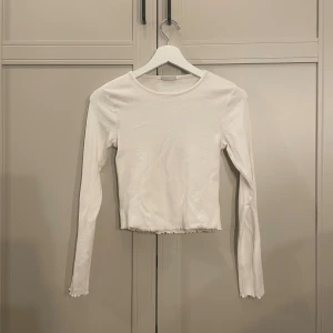 Vit långärmad topp från Kappahl - En stilren vit långärmad topp från Kappahl i ribbad bomull. Modellen är croppad med lätt vågig kant nertill och vid ärmslut, vilket ger en trendig touch. Perfekt att matcha med jeans eller kjol för en clean look.