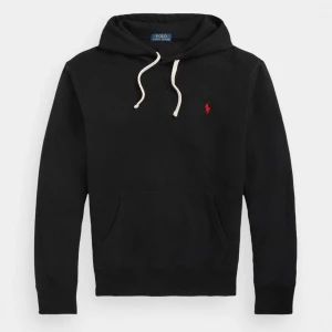Svart hoodie från Polo Ralph Lauren - Svart hoodie från Polo Ralph Lauren med klassisk röd logga broderad på bröstet. Tröjan har huva med vita snören och en stor magficka. Tillverkad i mjuk bomull och har långa ärmar. Perfekt för en avslappnad och stilren look.