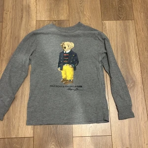 Grå långärmad tröja Polo Ralph Lauren - Grå långärmad tröja från Polo Ralph Lauren med ikoniskt Polo Bear-tryck framtill. Tröjan har rund halsringning och är tillverkad i mjuk bomull. Perfekt för dig som gillar klassisk stil med en twist. Super fin nu till hösten 