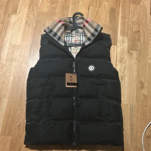 Svart dunväst från Burberry - Svart dunväst från Burberry med klassiskt rutigt foder i beige, svart, vitt och rött. Västen har dragkedja framtill, Burberry-logga på bröstet och hög krage. Perfekt för lager-på-lager och snygg streetstil. Kan även byta mot burberry puffer jacket men avtagningsbara armar!