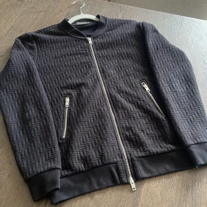 Svart quiltad tjocktröja med dragkedja från J.Lindeberg - Svart tjocktröja från J.Lindeberg med quiltat mönster och silverfärgade dragkedjor. Jackan har ribbade muddar vid ärmslut och nederkant, samt två fickor med dragkedja. 