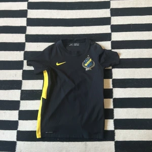 AIK 130 år Nike fotbollströja 147 - 158 CM. - AIK 130 år jubileumströja från Nike i svart med gula detaljer och klubbmärke på bröstet. Tillverkad i Nike Vaporknit-material som andas och har korta ärmar. Perfekt för dig som vill visa ditt AIK-stöd på match eller träning. Perfekt material, inga fläckar och inte en skråma. Knappt använd, i nyskick. Limited edition, jubileumströja. 