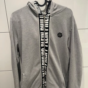 Grå hoodie från Philipp Plein med döskalle - Snygg grå hoodie från Philipp Plein med svart och vit logotypband längs dragkedjan och huvan. På ryggen finns ett stort tryck med döskalle och PP-loggor. Tröjan har huva, långa ärmar och fickor framtill. Perfekt för dig som gillar streetstyle och coola detaljer.