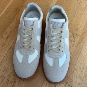 Maison Margiela sneakers i mocka och skinn - Snygga sneakers från Maison Margiela med vit skinnbas och ljusgrå mockadetaljer. Klassisk design med beige snörning och brun gummisula. Perfekta för dig som gillar stilrena och trendiga skor med en lyxig touch.