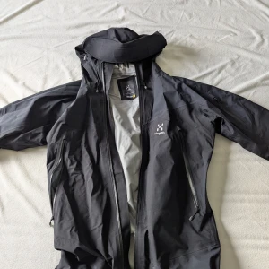 Svart Haglöfs regnjacka - Svart regnjacka från Haglöfs med justerbar huva, flera dragkedjeförsedda fickor och snygg minimalistisk design. Jackan är tillverkad i slitstark polyester och har långa ärmar samt hel dragkedja framtill. Perfekt för blåsiga dagar och outdoor-äventyr. Jackan är vattentätt, gortex 