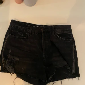 Svarta jeansshorts från Zara - Säljer ett par svarta jeansshorts från Zara med råa kanter och slitna detaljer både fram och bak. Shortsen har fem fickor, bälteshällor och klassisk knappgylf. Perfekta för en edgy och avslappnad look. De har glitter detaljer vid sidorna