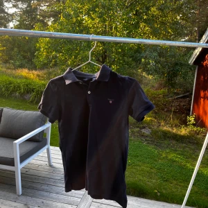 Mörkblå pikétröja från Gant - Säljer en svart piké från Gant med klassisk krage och knappar framtill. Tröjan har korta ärmar och en liten broderad logga på bröstet. Perfekt för en clean och stilren look. Det står 152 i stl men passade mig tills jag blev runt 164 ish.