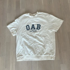 Vit t-shirt OAB authentic - Vit t-shirt från OAB med blått tryck 'OAB authentic' på bröstet. Klassisk rund hals och korta ärmar, tillverkad i mjuk bomull. Passar dig som gillar enkel och stilren streetwear med en touch av college-vibe.