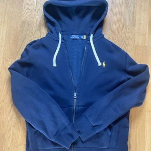 Marinblå hoodie från Polo Ralph Lauren - Snygg marinblå hoodie från Polo Ralph Lauren med gul broderad logga på bröstet. Tröjan har huva med vita snören, dragkedja framtill och två fickor. Klassisk och stilren design som alltid funkar.