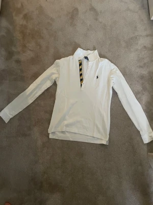 Vit pikétröja från Ralph Lauren Sport - Klassisk vit pikétröja från Ralph Lauren Sport i 100% bomull. Tröjan har lång ärm, krage och knäppning framtill med snygg detalj i gult och marinblått. Liten broderad logga på bröstet. Perfekt till en clean och sportig stil.