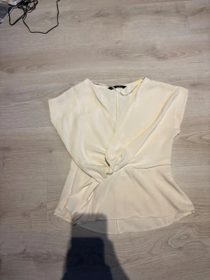 Vit omlottopp från Zara - Säljer en vit omlottopp från Zara i storlek S. Toppen har korta ärmar, v-ringning och knyt i sidan som ger en snygg siluett. Tillverkad i ett mjukt och lätt material som känns skönt mot huden. Perfekt att styla med jeans eller kjol.