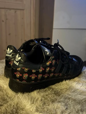 Svarta Adidas sneakers med färgglatt mönster - Vintage svarta Adidas sneakers tillverkat i 2007, i skinn med färgglatt mönster av små döskallar med sombreros på sidorna och hälen. Som representerar tradicionen dia de los muertos Klassiska tre ränder på sidan, svart sula och snörning. Adidas-logga i vitt på plös och häl. Perfekta för dig som vill sticka ut med coola detaljer.