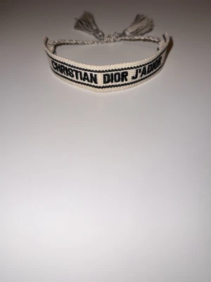 Christian Dior armband - Elegant och stilrent Christian Dior J’Adior-armband i klassisk design. Armbandet är tillverkat i vävd textil i en ljus, krämfärgad nyans med den ikoniska svarta texten “CHRISTIAN DIOR J’ADIOR” framtill. Justerbar knytning med dekorativa tofsar som ger både komfort och en exklusiv känsla.