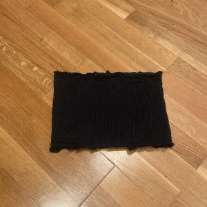 Svart bandeau tubtopp från SOC - Säljer en svart bandeau tubtopp från SOC i storlek XS/S. Toppen är smockad med stretchigt tyg och har små volangdetaljer upptill och nedtill. Perfekt till sommaren och enkel att styla med olika outfits.