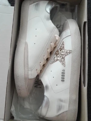 Golden Goose sneakers med glitterstjärna - Snygga vita sneakers från Golden Goose med silvriga detaljer och en glittrig stjärna på sidan. Skorna har snörning, platt sula och inslag av mocka vid tån och hälen. Perfekta för dig som vill sticka ut med en cool och trendig look.