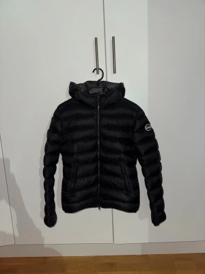 Svart dunjacka från Colmar, strl 44 - Säljer en svart dunjacka från Colmar med huva och dragkedja framtill. Jackan har en quiltad design, två fickor med dragkedja och en snygg logga på ärmen. Perfekt för kalla dagar och riktigt skön att ha på sig.