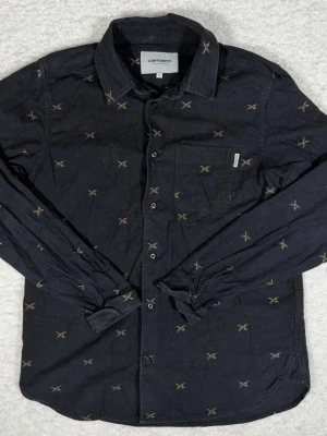 Svart Carhartt skjorta med mönster - Svart skjorta från Carhartt Work In Progress i storlek S. Skjortan har ett brunt broderat mönster över hela plagget, klassisk krage, knappstängning och en bröstficka med Carhartt-tag. Långärmad och tillverkad i mjuk bomull.