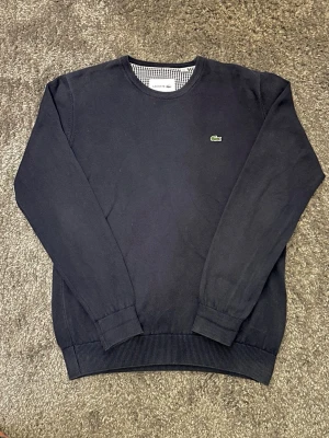 Lacoste tröja - Snygg mörkblå stickad tröja från Lacoste med klassisk rund hals och det ikoniska krokodilmärket broderat på bröstet. Tröjan har långa ärmar och ribbade muddar vid ärmslut och nederkant. Perfekt för dig som gillar stilrena och tidlösa plagg. 