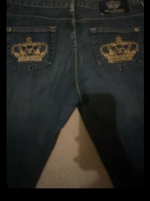 Mörkblå jeans med guldbroderade kronor - Säljer ett par mörkblå jeans med raka ben och snygga guldbroderade kronor på bakfickorna. Jeansen har klassisk femficksdesign och är tillverkade i slitstarkt denimtyg. Perfekta för dig som vill sticka ut med coola detaljer! OBS tipsar till dig som är lite kortare, tex 155-162!!