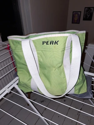 Grön shoppingväska Peak Performance - Snygg grön shoppingväska från Peak Performance med vita detaljer och breda handtag. Väskan är rymlig och har en vit insida samt en innerficka. Perfekt för att bära med sig allt man behöver under dagen. Stängs med dragsko. Djur finns i hemmet