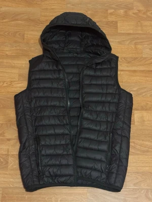 Svart dunväst med huva - Säljer en svart dunväst med huva och dragkedja framtill. Västen har quiltad design och två fickor med dragkedja. Perfekt att slänga över en hoodie för extra värme under kyliga dagar.
