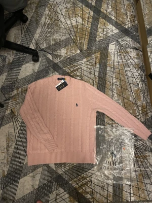 Ljusrosa stickad tröja Polo Ralph Lauren - Supersnygg ljusrosa stickad tröja från Polo Ralph Lauren i klassisk kabelstickad design. Tröjan har rund hals, långa ärmar och den ikoniska loggan broderad på bröstet. Perfekt till jeans eller kjol för en clean och fräsch look.