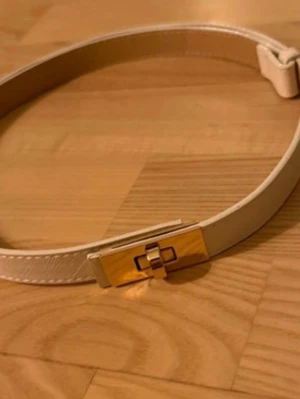 Beige bälte med guldfärgat spänne - Snyggt beige bälte med ett stilrent guldfärgat spänne som har en unik design. Bältet är smalt och passar perfekt till jeans eller klänning för att ge outfiten en extra touch. Materialet är slätt och har diskreta sömmar längs kanterna.