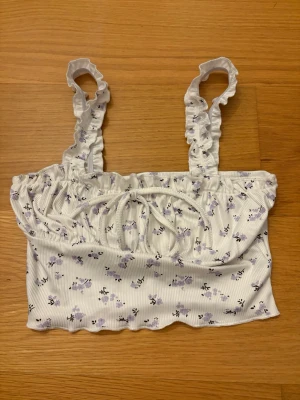 Blommig vit croppad topp från SHEIN, storlek XS - Supersöt croppad topp från SHEIN i storlek XS i vit med ljuslila blommigt mönster. Toppen har volangkantade axelband, ribbad struktur och en söt knytning framtill. Perfekt för sommarens alla häng! Materialet är mjukt och stretchigt. 95% Polyester, 5% Elastan.
