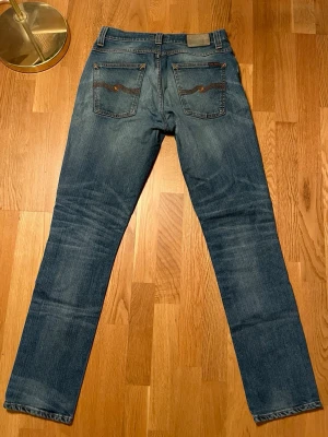 Blå Nudie Jeans, storlek 32/34 - Säljer ett par klassiska blå jeans från Nudie Jeans i modellen East Coast Blues. Jeansen har raka ben, fem fickor och snygga detaljer med orange sömmar och broderade bakfickor. Materialet är 98% ekologisk bomull och 2% elastan för extra komfort.