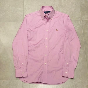 Raffe skjorta - Säljer en slim fit skjorta från Ralph Lauren i ljusrosa bomull. Klassisk button-down krage, broderad logga på bröstet och vita knappar framtill. Skjortan har lång ärm och stilren design som ger en fräsch vibe. Perfekt för dig som gillar snygga och tidlösa plagg.