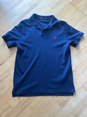 Marinblå piké från Polo Ralph Lauren - Säljer en marinblå pikétröja från Polo Ralph Lauren i custom slim fit. Klassisk krage, två knappar och den ikoniska röda broderade loggan på bröstet. Tillverkad i mjuk bomull som känns skön mot huden. Perfekt för en clean och stilren look. Priset är förhandlingsbart!
