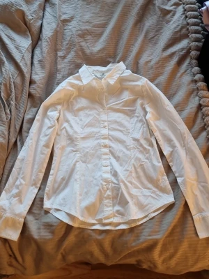 Vit klassisk skjorta med knappar - Säljer en stilren vit skjorta från H&M med klassisk krage och långa ärmar. Skjortan har en figurnära passform och knäppning framtill.