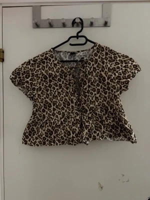 Leopardmönstrad blus med snörning - Säljer en kortärmad blus med leopardmönster och snörning framtill. Toppen har en lös passform och volang nedtill, vilket ger en avslappnad och trendig look. Perfekt för dig som vill sticka ut lite extra. Materialet känns lätt och luftigt.