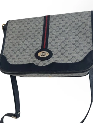 Gucci axelväska med monogram - Säljer en klassisk Gucci axelväska i grått och mörkblått med GG-monogram över hela väskan. Väskan har en justerbar axelrem, detaljer i skinn och en ikonisk röd-blå rand på framsidan. Stängs med ett metallspänne med GG-logga och har flera fack inuti. En vintage modell som är riktigt svår att få tag i, den har dock defekter därav priset