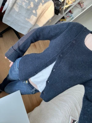 Mörkgrå stickad kofta med knappar - Mysig mörkgrå stickad kofta med rund hals och knäppning framtill. Koftan har långa ärmar och ribbade muddar vid ärmslut och nederkant. Perfekt att slänga över en t-shirt för en chill look. Ny pris 300kr, bara använd 1 gång