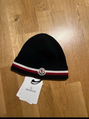Blå  Moncler mössa med röd/vit rand - Snygg svart Moncler mössa med klassisk röd och vit rand runt kanten och Moncler-logga framtill. Mössan är ribbstickad och kommer med originalpåse och taggar. Perfekt accessoar för dig som vill ha en stilren och exklusiv look.