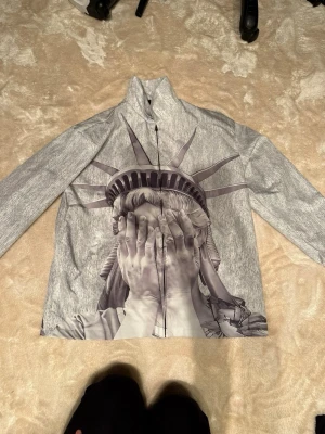 Grå vindjacka med Frihetsgudinnan-print - Säljer en grå vindjacka med ett stort, detaljerat print av Frihetsgudinnan som täcker både fram- och baksidan. Jackan har dragkedja framtill och lång ärm. Materialet känns lätt och passar perfekt för dig som vill sticka ut med en unik design.