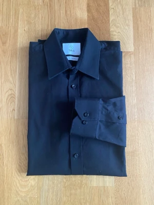 Svart skjorta från Bläck, Tailored Fit - Klassisk svart skjorta från Bläck med lång ärm och skräddarsydd passform. Skjortan har en stilren krage och knappar framtill.  Perfekt för dig som gillar en clean och enkel look.