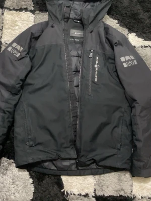 Svart Sail Racing vindjacka med huva - Säljer en svart vindjacka från Sail Racing i storlek S. Jackan har huva, dragkedja framtill och en bröstficka med logga. Materialet är slitstark polyester och jackan har långärmade ärmar med tryck på båda. Perfekt för dig som gillar stilren och sportig look.