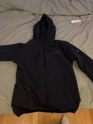 Peak Performance Jacka - Säljer en svart vindjacka från Peak Performance, modell Eddie Jacket. Jackan har huva med snörning, två fickor med dragkedja och diskret logga på bröstet. Perfekt för blåsiga dagar och enkel att matcha med allt.