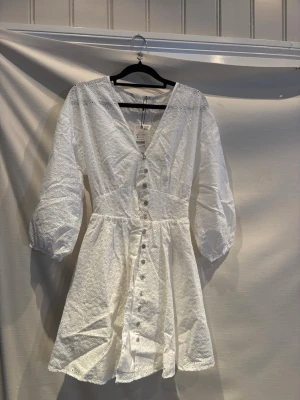 Vit kort klänning med broderade blommor - Supersöt vit kort klänning i bomull med broderade blommor, v-ringning och knappar framtill. Klänningen har trekvartsärm med lätt puff och resår i midjan för en snygg siluett. Perfekt för sommarens alla tillfällen!