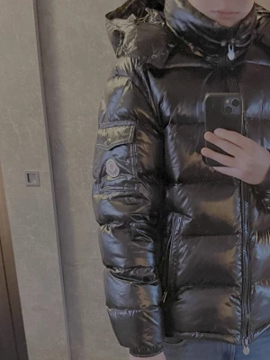 Moncler maya - Svart glansig pufferjacka från Moncler med huva och dragkedja framtill. Jackan har två fickor med dragkedja och Moncler-logga på ärmen. Den är quiltad och har en riktigt fet streetstil som passar perfekt när det är kallt ute. Använd i 2 dgr, kvitto finns!