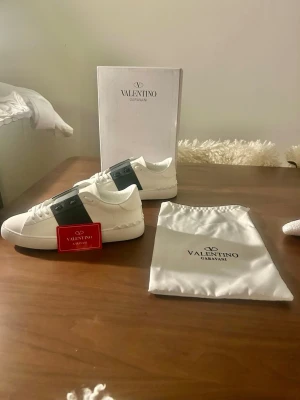 Valentino Garavani Open sneakers vit/svart - Säljer ett par Valentino Garavani Open sneakers i vitt skinn med svart band över snörningen och ikoniska nitar runt hälen. Klassisk rund tå och platt sula. Kommer med originalkartong och dustbag. Perfekta för dig som vill ha en clean men lyxig look.