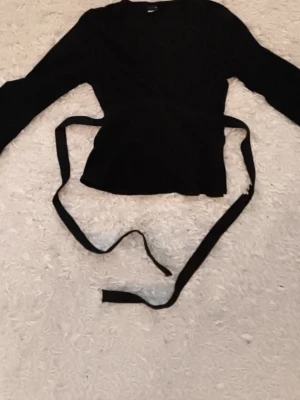 Svart topp från Gina Tricot - Säljer en svart v-ringad blus från Gina Tricot med långa ärmar och knytband i midjan. Toppen har en v-ringad skärning och är tillverkad i ett mjukt material som ger en skön känsla. Perfekt att styla med jeans eller kjol.