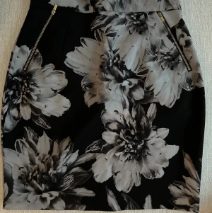 Blommig svartvit kjol från H&M - Säljer en svartvit kortkjol från H&M med stora blommönster och snygga guldfärgade dragkedjor fram och bak. Kjolen har en rak passform och är gjord i ett mjukt polyestertyg som känns skönt mot huden. Perfekt för dig som vill sticka ut lite extra!