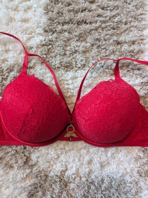 Röd spets-bh med guldring - Snygg röd bh med vadderade kupor och spetsdetaljer. Dekorativ guldfärgad ring framtill mellan kuporna och dubbla axelband för extra stil. Perfekt för dig som vill ha något extra under kläderna.